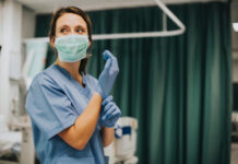 Nurse wearing scrubs and mask putting on gloves.