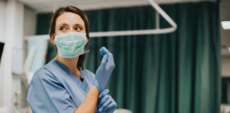 Nurse wearing scrubs and mask putting on gloves.