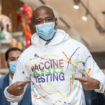 Man wearing Vaccine and Testing hoodie, pointing at text.