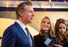 Airport SHOWDOWN: Newsom Dares Trump… A man in a suit speaking at an event with two women nearby, one smiling and the other holding a microphone