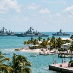 A fleet of naval ships sailing in the ocean near a tropical shore
