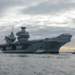 China’s Fleet Grows—Protection Lags Aircraft carrier with tugboats on a cloudy sea.