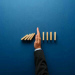 Hand stopping dominoes from falling on blue background.