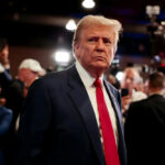 Missile Plot Whispered—Who Lied To Trump? A serious-looking man in a suit with a red tie surrounded by a crowd and cameras