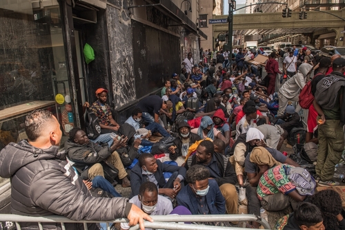 A large group of people sitting on a city street, some wearing masks