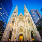 Gothic cathedral illuminated at dusk surrounded by modern skyscrapers