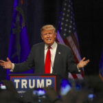 A speaker gesturing passionately at a political rally with American flags in the background
