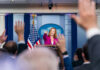 Press secretary at podium, answering reporters questions, raised hands.